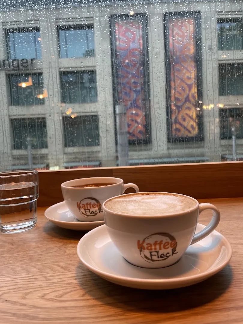 Zwei Cappuccino-Tassen mit Kaffee-Fleck-Logo am Fensterplatz